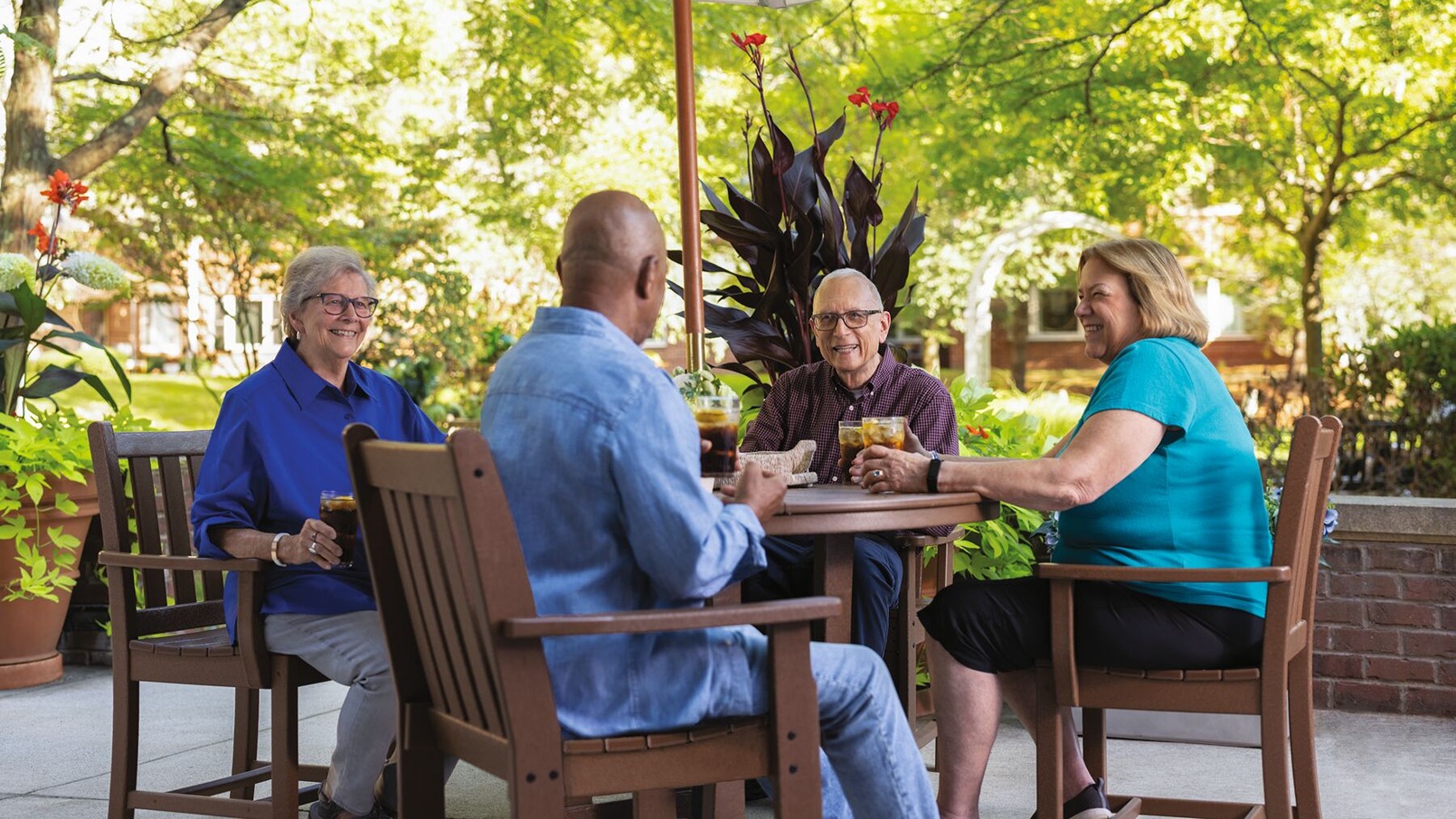 A multicultural group of four Erickson residents gather at a patio table laughing and talking. 