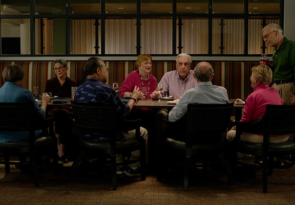 Large group of seniors gathered in a cozy, welcoming dining area at a residential community, enjoying conversation, drinks and meals together.