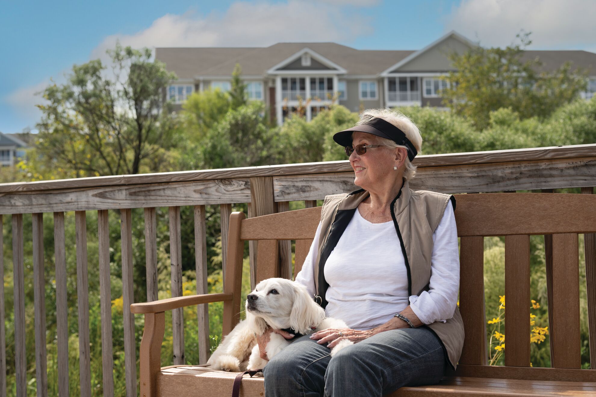 A woman and her dog sit on a bench outside. Green trees and a residence building are in the background.
