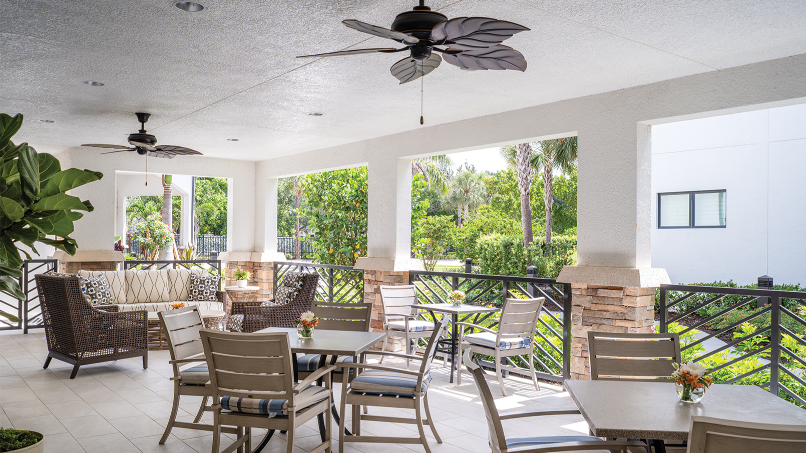 Covered outdoor patio with ceiling fans, wicker lounge seating, and dining tables overlooking a landscaped garden with palm trees and greenery.