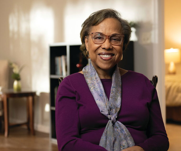 Smiling older woman wearing glasses, a purple top, and a light patterned scarf, seated in a warmly lit living room with a bookcase and bedroom in the background.