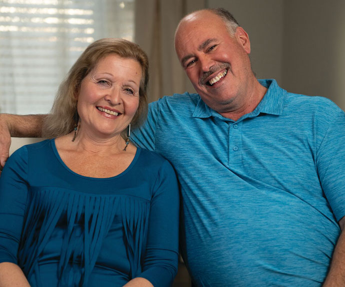 Smiling older couple sitting close together indoors, both wearing blue tops, with the man’s arm around the woman.