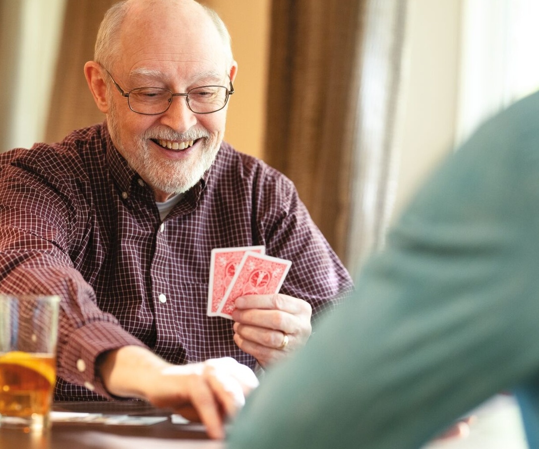 Older man smiling while playing cards at a table with a drink, interacting with another person.