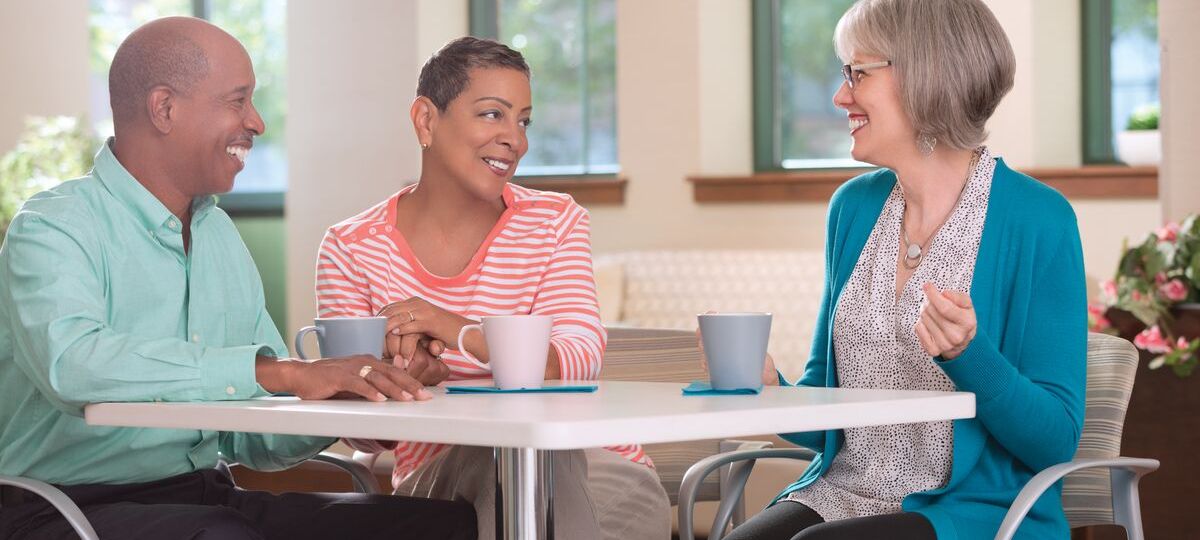 Three seniors sit at a table with coffee laughing and talking