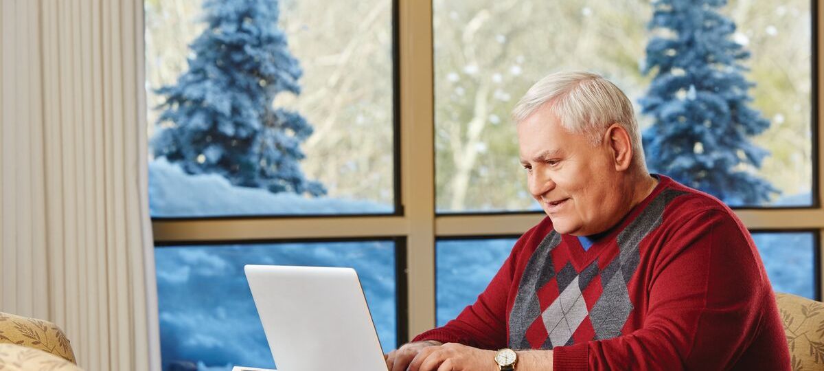 Man sits at a table with a laptop. He is indoors but there is snow outside.
