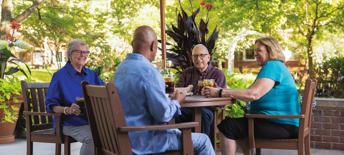A multicultural group of four Erickson residents gather at a patio table laughing and talking. 