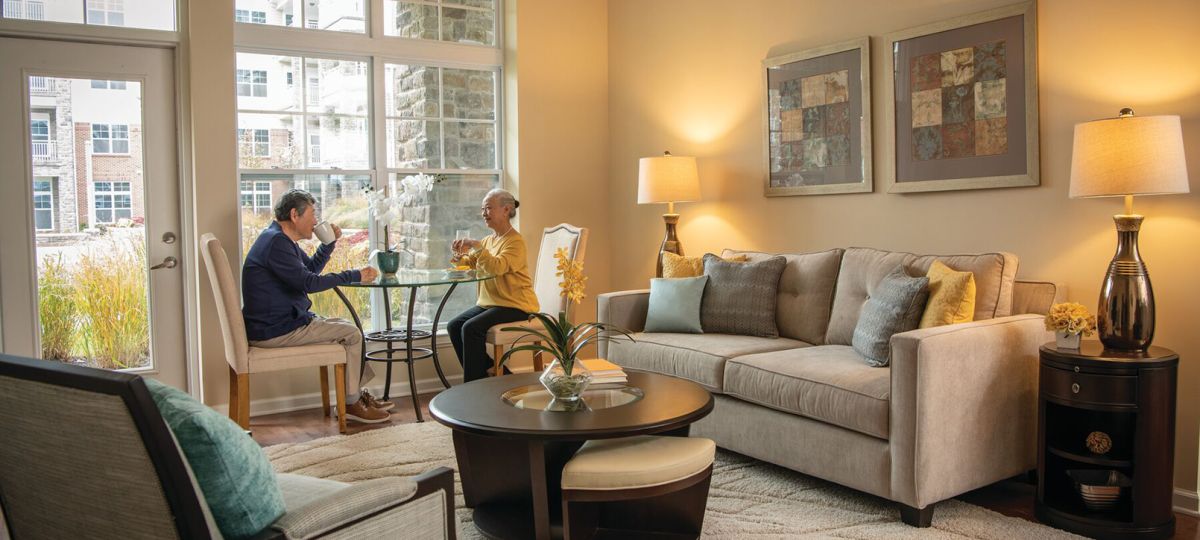 Bright living room with large windows overlooking a courtyard in an apartment community. A couple sits at a small table by the window enjoying a meal together.