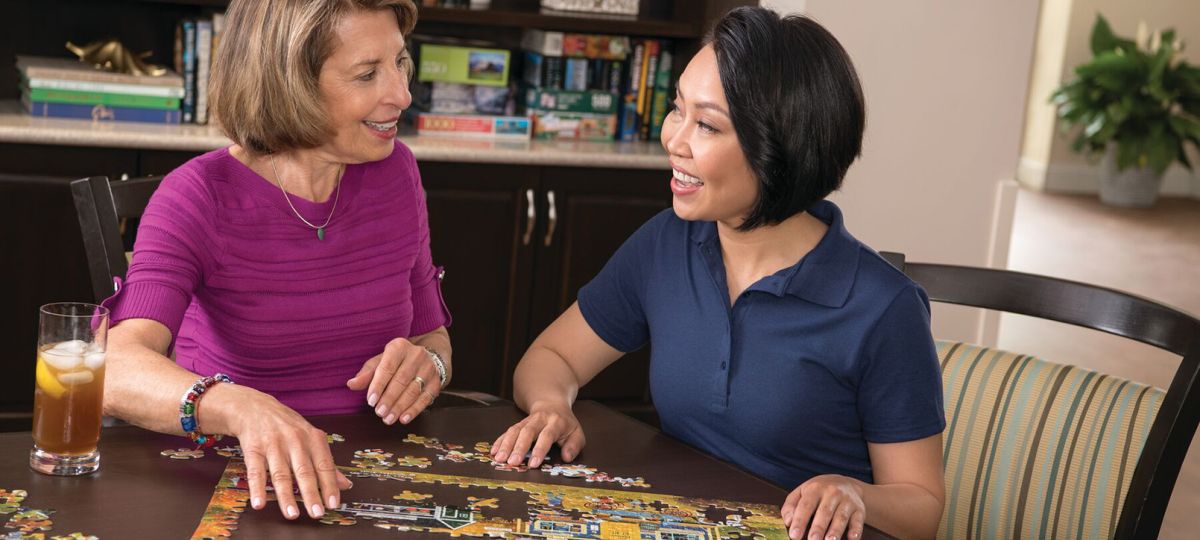 A cheery Erickson employee puts together a puzzle with a smiling resident in a community multipurpose room.