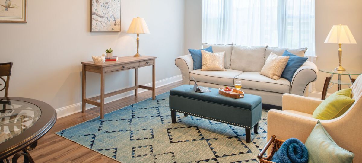 A bright and airy senior apartment living room with a large window, comfortable seating arranged for conversation, and tasteful decor creating a warm, inviting space.
