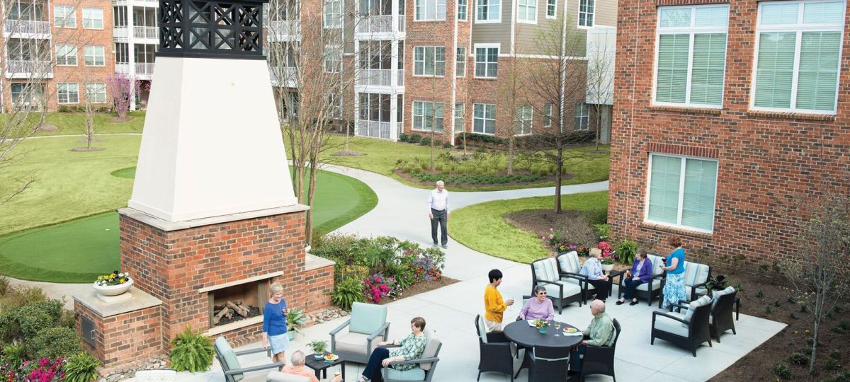 Open inviting outdoor patio with comfortable seating, shade umbrellas, and potted plants. Senior residents relax and socialize in a peaceful garden setting.
