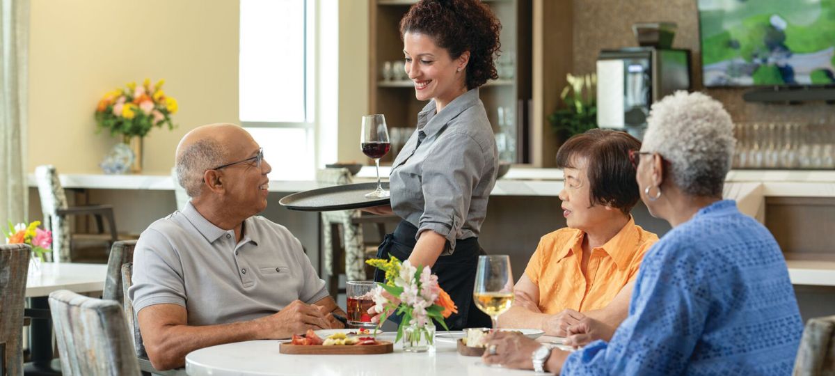 Server bringing meals to residents seated at a dining table in an Erickson Senior Living dining facility.