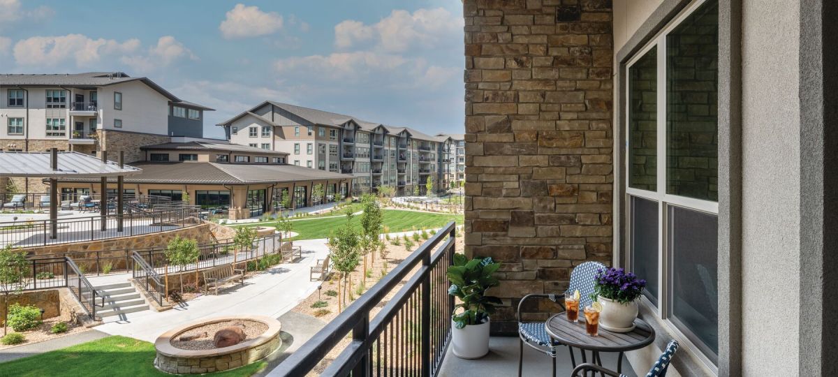 Spacious balcony at a senior living community apartment overlooking a beautifully landscaped courtyard with lush greenery, walking paths, and seating areas.