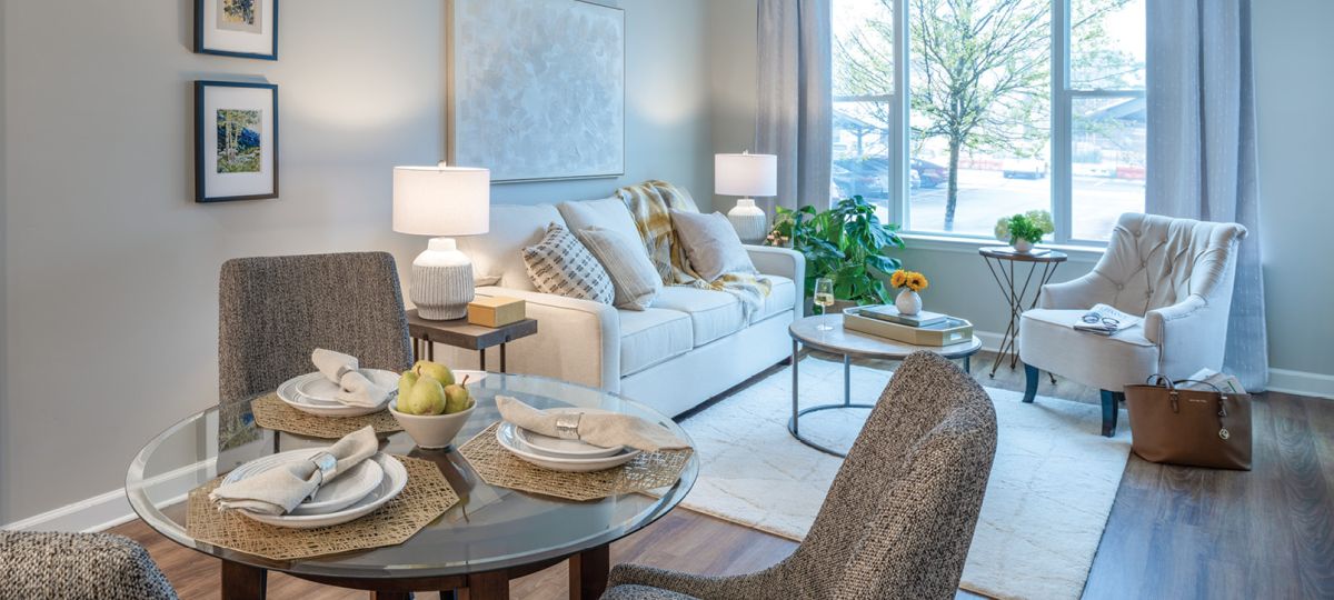 A stylish, living room with modern vinyl flooring and featuring a couch with decorative pillows and a small circular breakfast table with four cushy chairs. A cozy armchair is positioned near a large window with a scenic view of greenery.