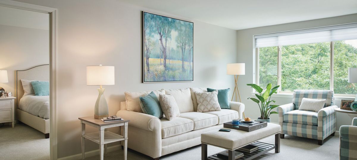 A bright and airy living room with a cream-colored sofa adorned with decorative pillows, a checkered armchair, and a large window with a lush green outdoor view. A soft ottoman and wooden coffee table add functionality, while a floor lamp and potted plant bring warmth and freshness. The adjacent bedroom, visible through an open doorway, features a cozy bed with light blue accents.