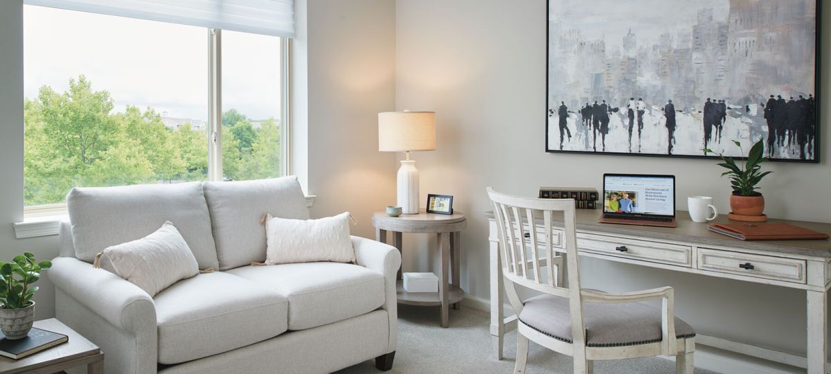 A cozy home office and sitting area with a light-colored loveseat adorned with soft pillows, positioned by a large window overlooking lush greenery. A rustic wooden desk with a distressed finish holds a laptop, books, and a potted plant, accompanied by a matching chair with a cushioned seat.