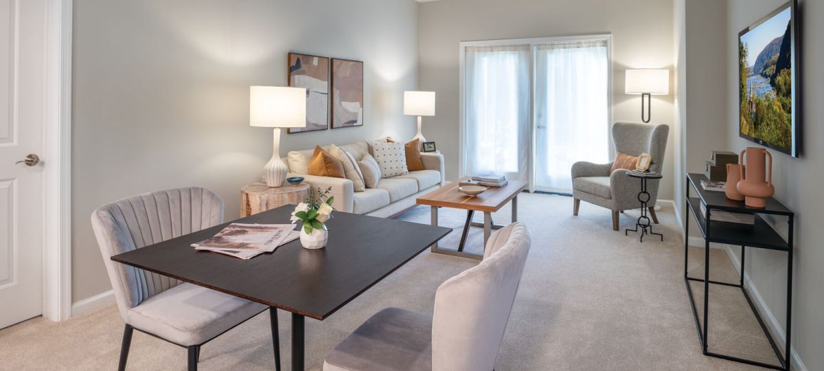 Modern living and dining area with neutral tones, featuring a square dining table with plush chairs, a cozy beige sofa, contemporary wall art, and a TV mounted on the wall.
