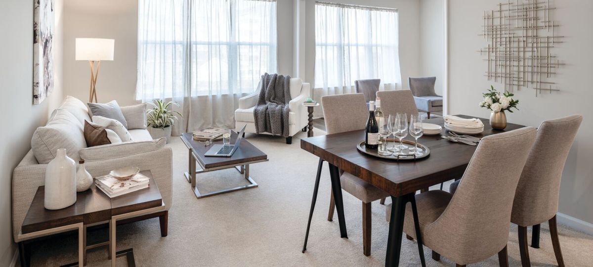 Elegant open-concept living and dining room with large windows, beige carpet, a light gray sectional sofa, a dark wood dining table with cushioned chairs, and modern decor elements.