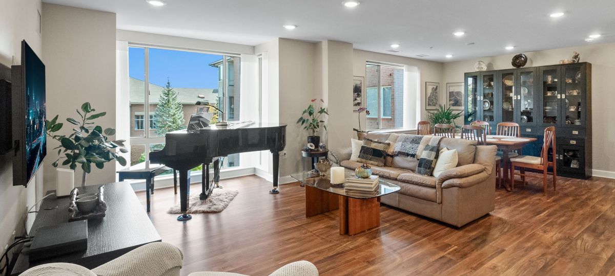 Spacious, sunlit living and dining area in an Erickson Senior Living apartment, featuring a grand piano by large windows, a cozy seating arrangement, hardwood floors, and a formal dining set with a glass-front china cabinet.