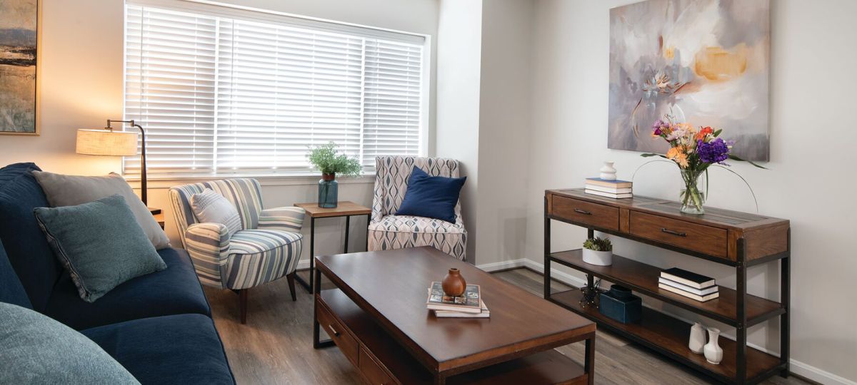 A modern living room with a navy blue sofa, striped and patterned accent chairs, a wooden coffee table, and a console table decorated with books, vases, and a flower arrangement. Large windows with white blinds allow natural light to brighten the space, which features neutral tones and contemporary artwork on the wall.