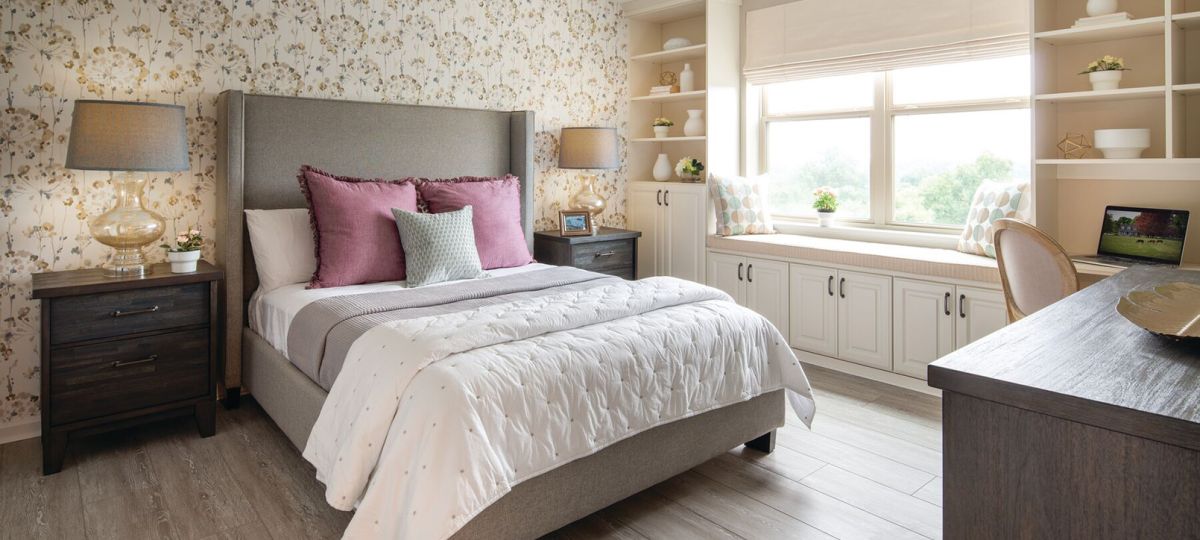 Elegant bedroom with a gray upholstered bed, white bedding, mauve and sage pillows, floral wallpaper, and dark wood nightstands. A built-in window seat with storage and a sleek desk add charm and functionality.