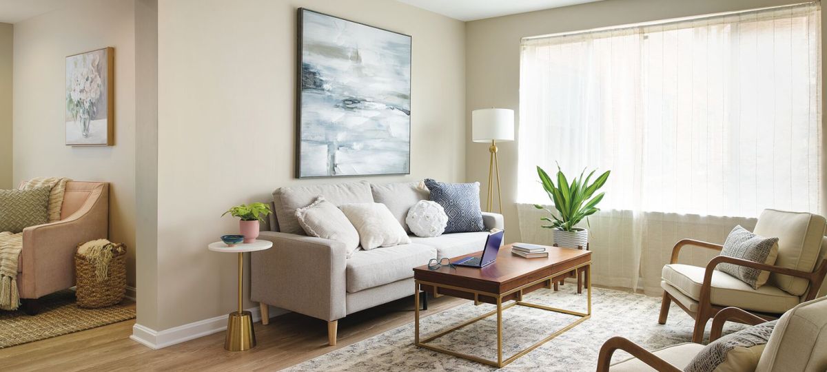 Bright and modern living room with neutral tones, featuring a beige sofa, wooden coffee table, accent chairs, and a large window with sheer curtains.