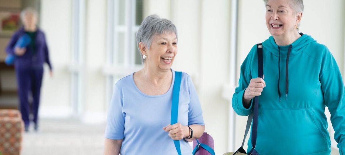 Two senior women smiling and walking together indoors, each carrying a yoga mat, dressed in casual workout clothes, with a third woman in the background also holding a yoga mat.