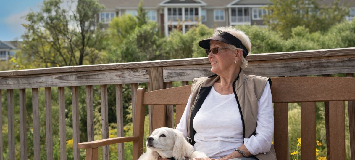A woman and her dog sit on a bench outside. Green trees and a residence building are in the background.