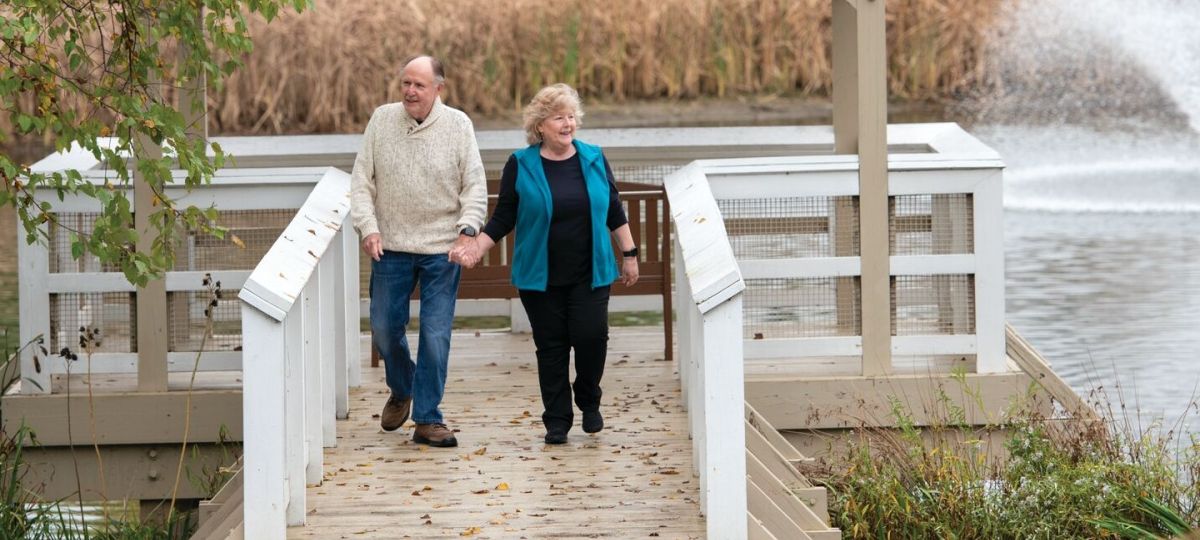 Senior couple holding hands and walking together on a wooden dock by a pond, surrounded by autumn foliage and reeds, with a fountain in the background.