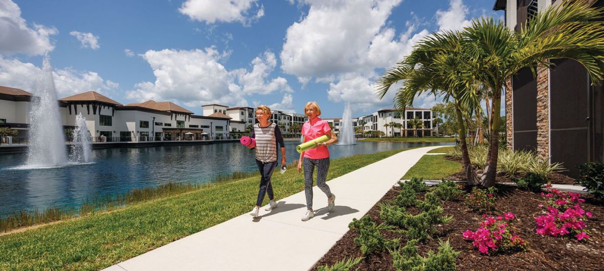 Couple strolls along a scenic paved path holding yoga mats beside a lake with a fountain at Siena Lakes, an Erickson Senior Living community.