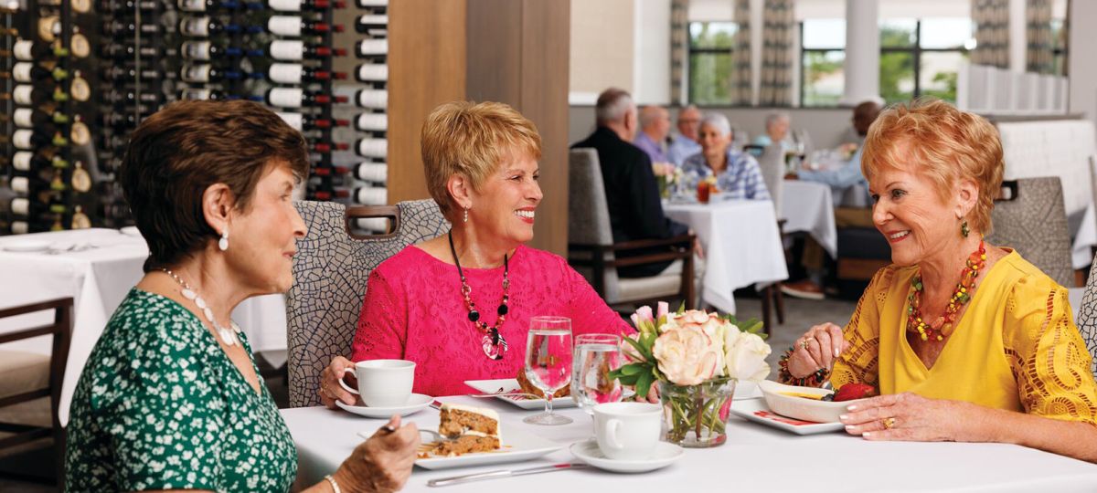 Three older women enjoy coffee and dessert together at a white tablecloth restaurant with a wine wall backdrop.