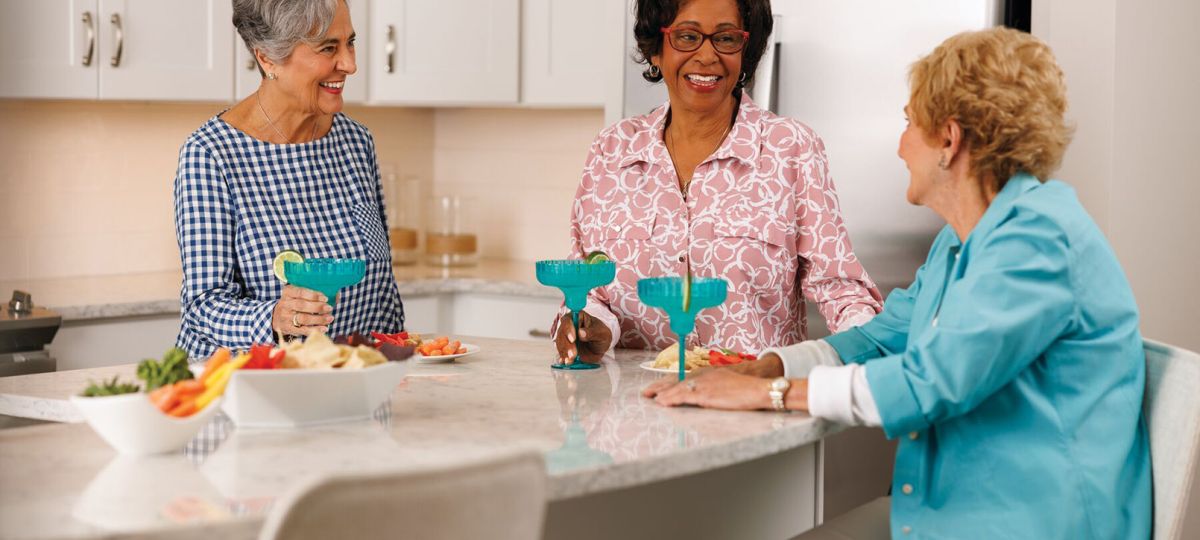 Three women share laughter and conversation in a bright, modern Erickson Senior Living apartment kitchen.