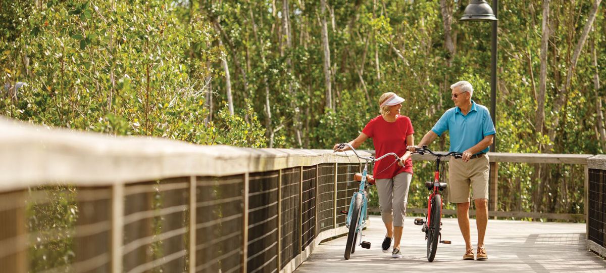 A couple walks side by side with their bikes along a wooded trail, enjoying a conversation.