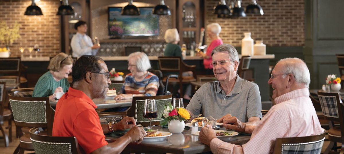 Three senior men smiling and dining together in a well-lit, modern retirement community restaurant, with other seniors socializing in the background.