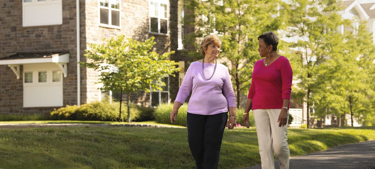 Two women walking outside on a walking path talking to each other. A residence building and trees are in the background.