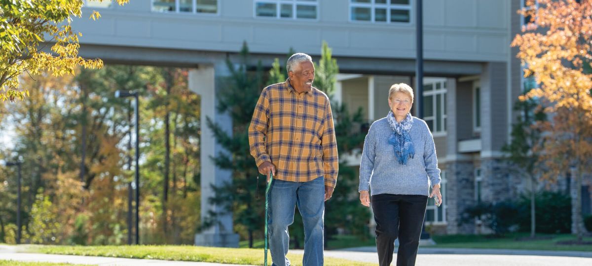 A man and a woman walking down the sidewalk smiling. A residence building's walkway features in the background
