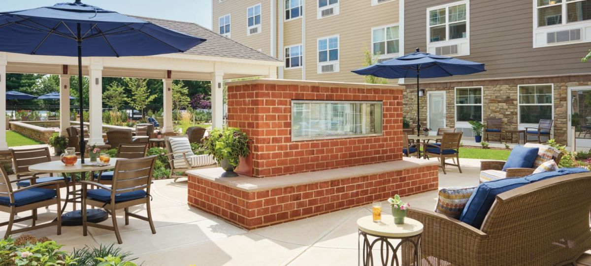 Outdoor courtyard with patio seating, blue umbrellas, garden landscaping, and a central brick fireplace feature.