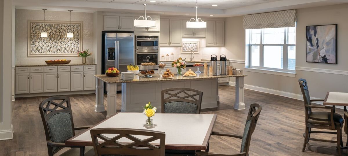 Spacious communal kitchen and dining area with a large island set with pastries, fruit, and coffee service, surrounded by light-colored cabinetry, stainless steel appliances, and multiple seating tables.