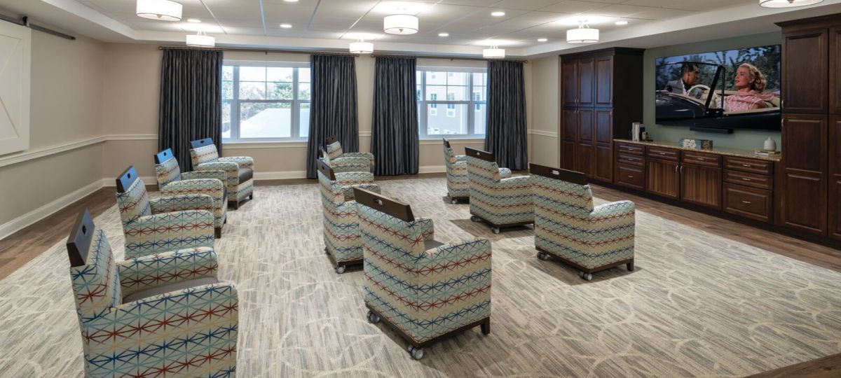 Theater-style lounge with patterned armchairs facing a large wall‑mounted TV and built‑in wooden cabinetry.
