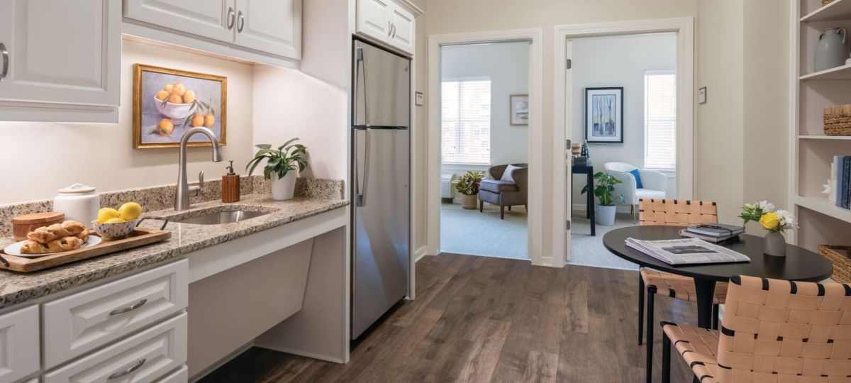 Compact kitchenette with granite countertops, white cabinets, and a small dining table, leading to two adjoining rooms with seating and decor.