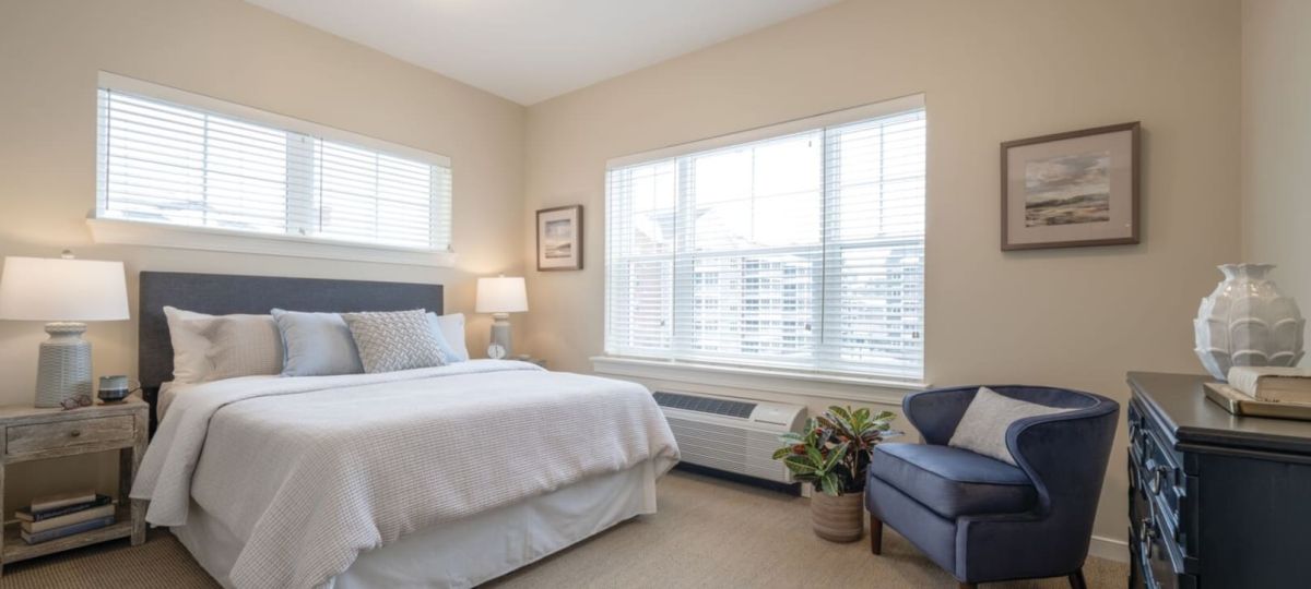 Bright bedroom with neatly made bed, armchair, dresser, and large windows.
