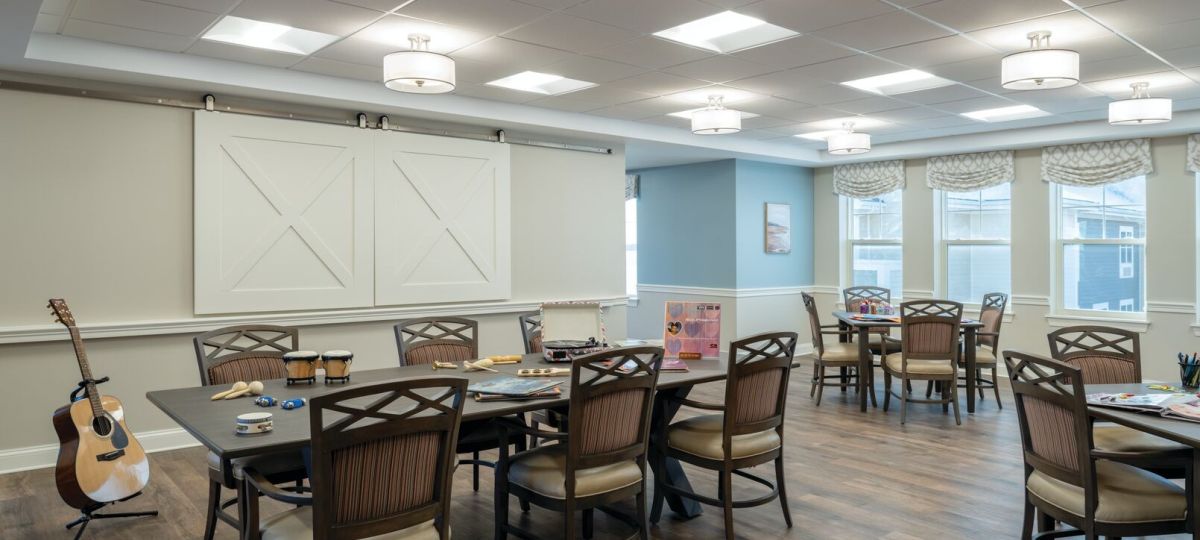 Creative activity room with multiple tables, art supplies, musical instruments, and large windows along one wall.