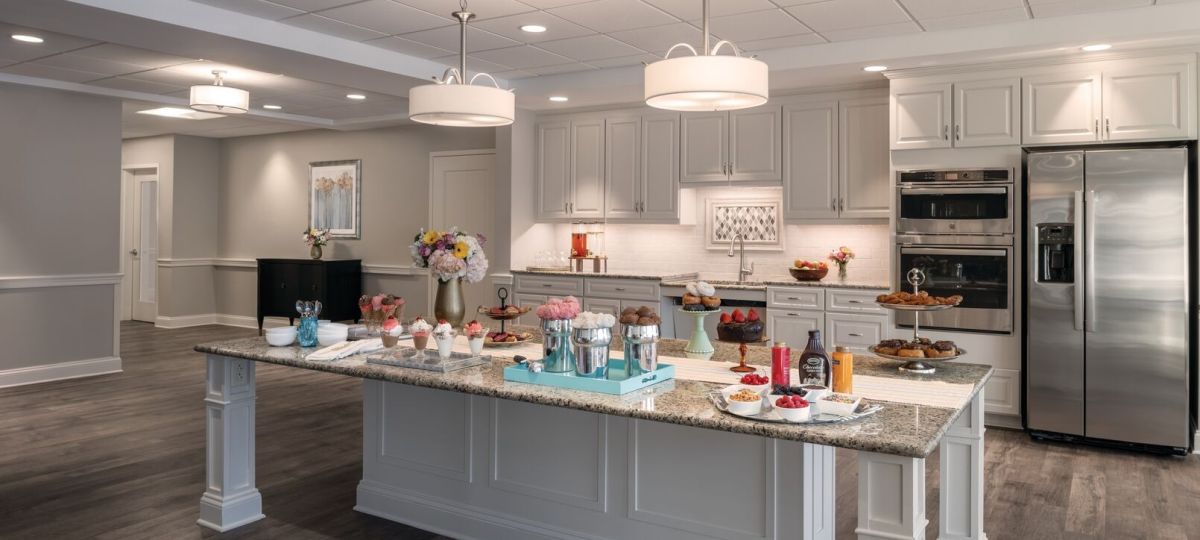 Spacious community kitchen with a large island set with desserts and beverages, white cabinetry, and stainless steel appliances.