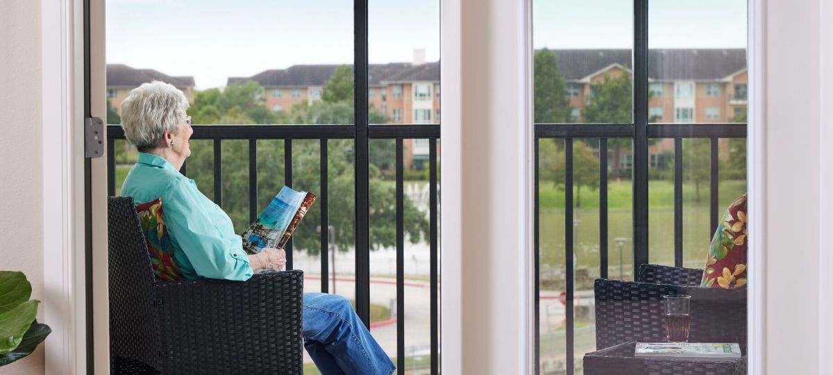 A woman sits in a sunroom, looking out residence buildings across a green field.
