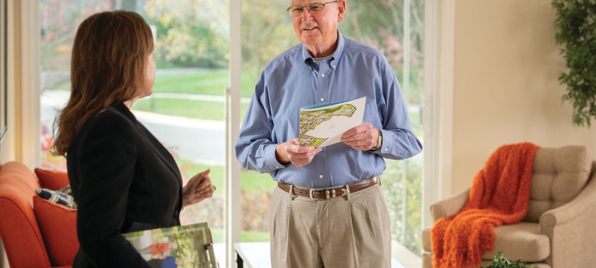 A man and woman stand in a model apartment living room talking. The man is holding a brochure.