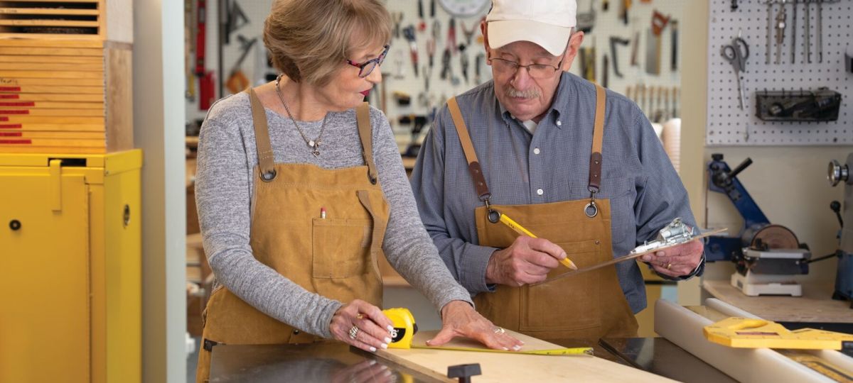 Older couple in a workshop measuring wood together with tools and aprons.