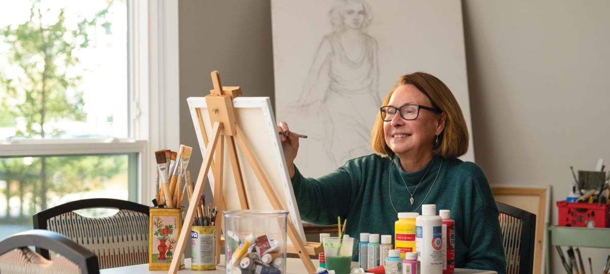 Older woman painting on a canvas at an easel, surrounded by art supplies in a bright, creative studio space. 