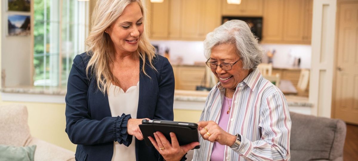 An Erickson Senior Living staff member assisting an older woman with a tablet in a comfortable home setting, smiling as they learn and use technology together.