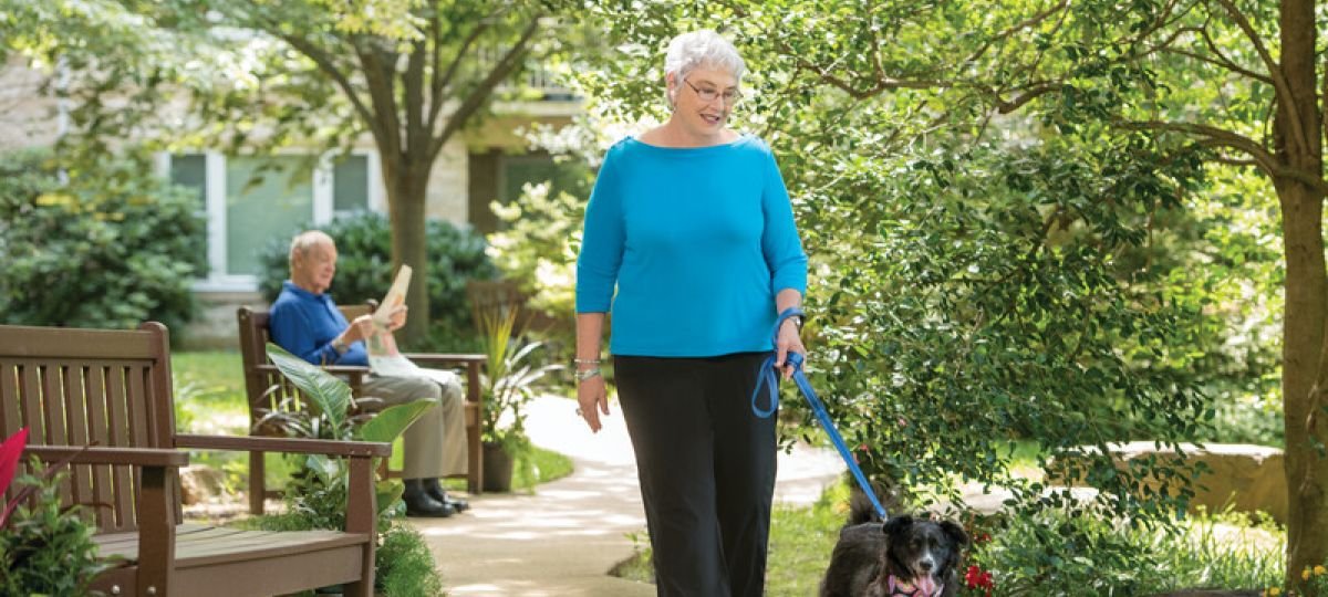 Older woman walking a dog along a garden path in an Erickson Senior Living community courtyard, with another resident seated on a bench reading nearby.