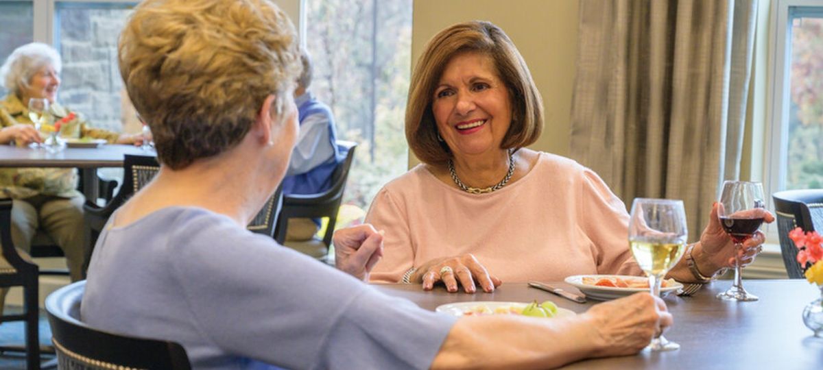 Two older women smiling and talking over a meal with drinks in a bright senior living dining room, with other residents seated in the background.