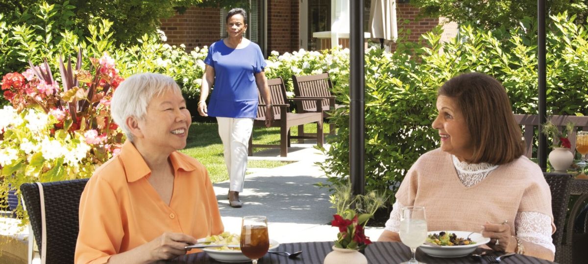 Two women enjoying lunch and conversation at an outdoor patio table surrounded by greenery.
